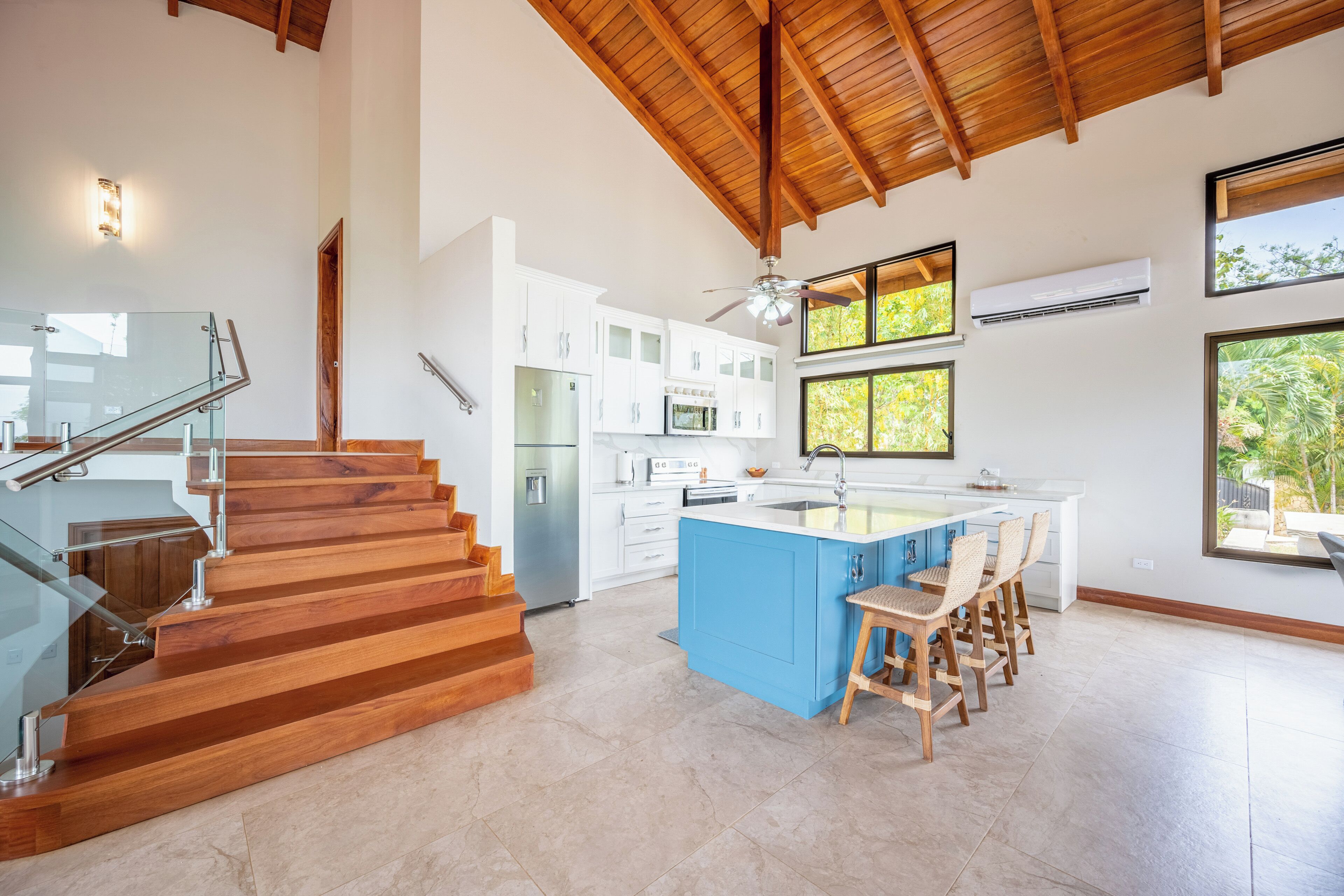 A modern kitchen with a central island, wooden staircase, and large windows.