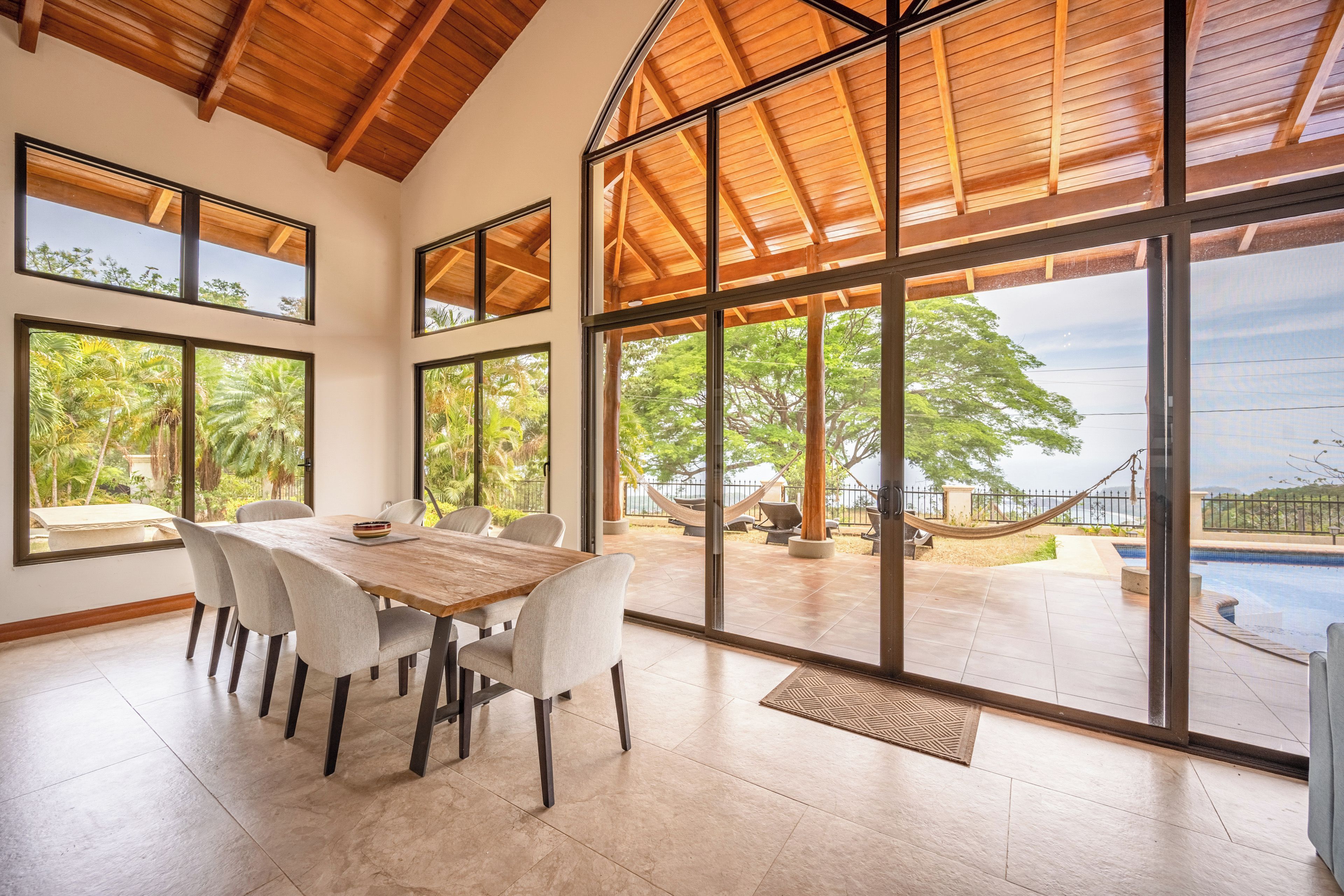 A spacious dining area with a large table and chairs, large windows offering views of a pool and outdoor seating.