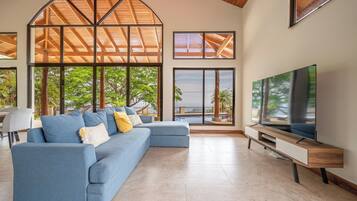 A modern living room with a sectional sofa, a flat-screen TV, and large windows offering a view of the outdoors.