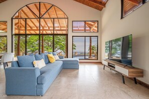 A modern living room with a sectional sofa, a flat-screen TV, and large windows offering a view of the outdoors.