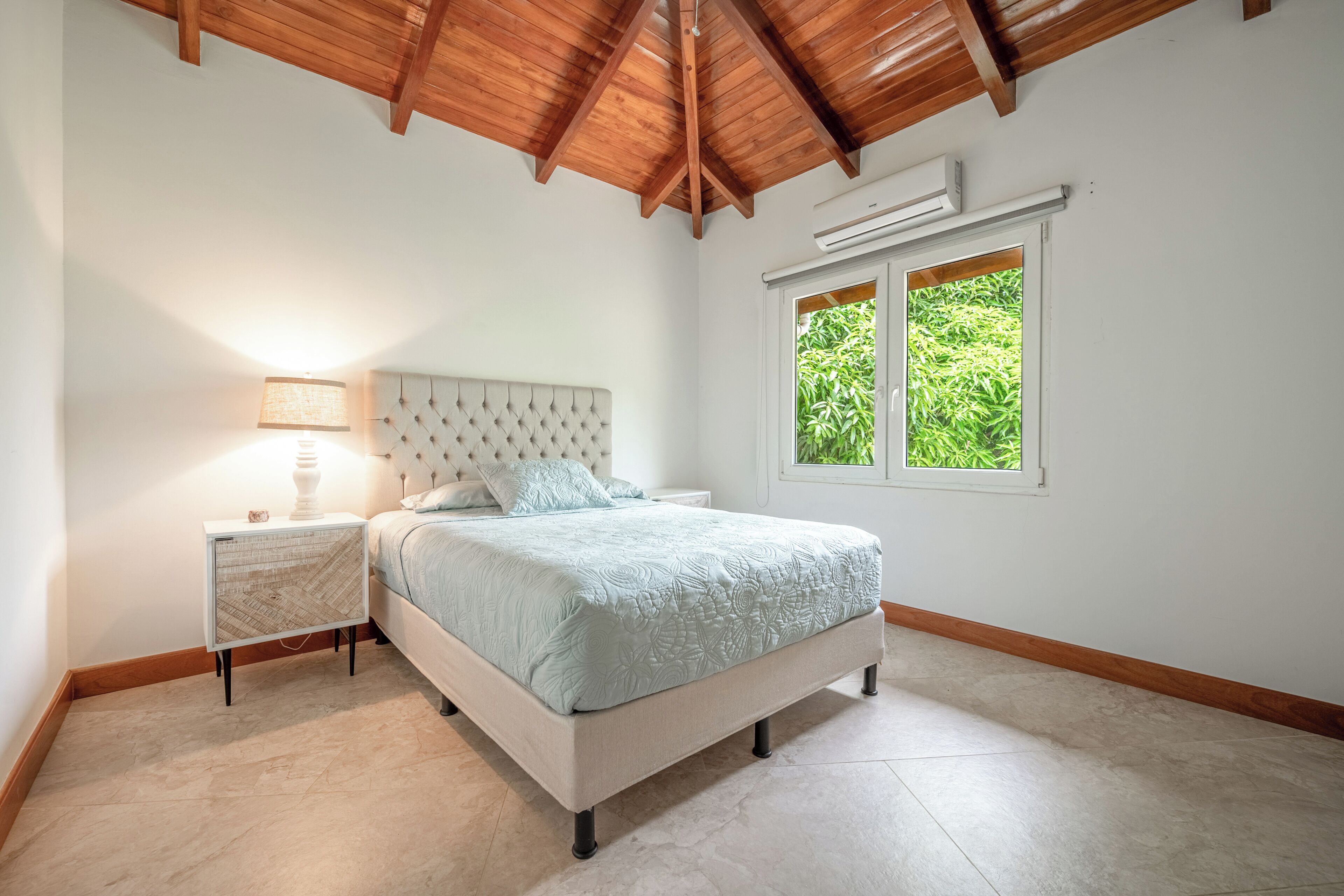 A bedroom with a bed, nightstand, lamp, and a window with a view of greenery.