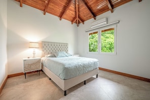 A bedroom with a bed, nightstand, lamp, and a window with a view of greenery.