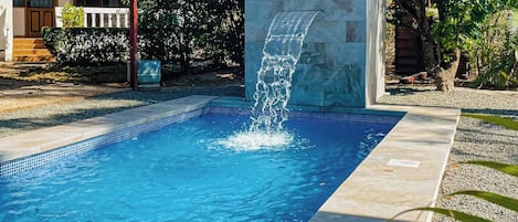 Zwembad met waterval, omringd door een stenen muur en een overdekt gebied met een trap naar een deur.