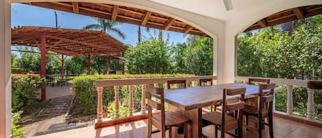 A covered outdoor dining area with a table and chairs, a ceiling fan, and a view of a garden with various plants and flowers.