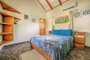 A bedroom with a bed, wooden headboard, stone floor, and a window with a view of greenery.