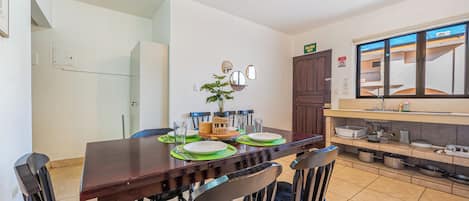 Un comedor con una mesa de madera dispuesta para cuatro, sillas, un ventilador de techo y una cocina con un lavabo y una encimera a la vista.