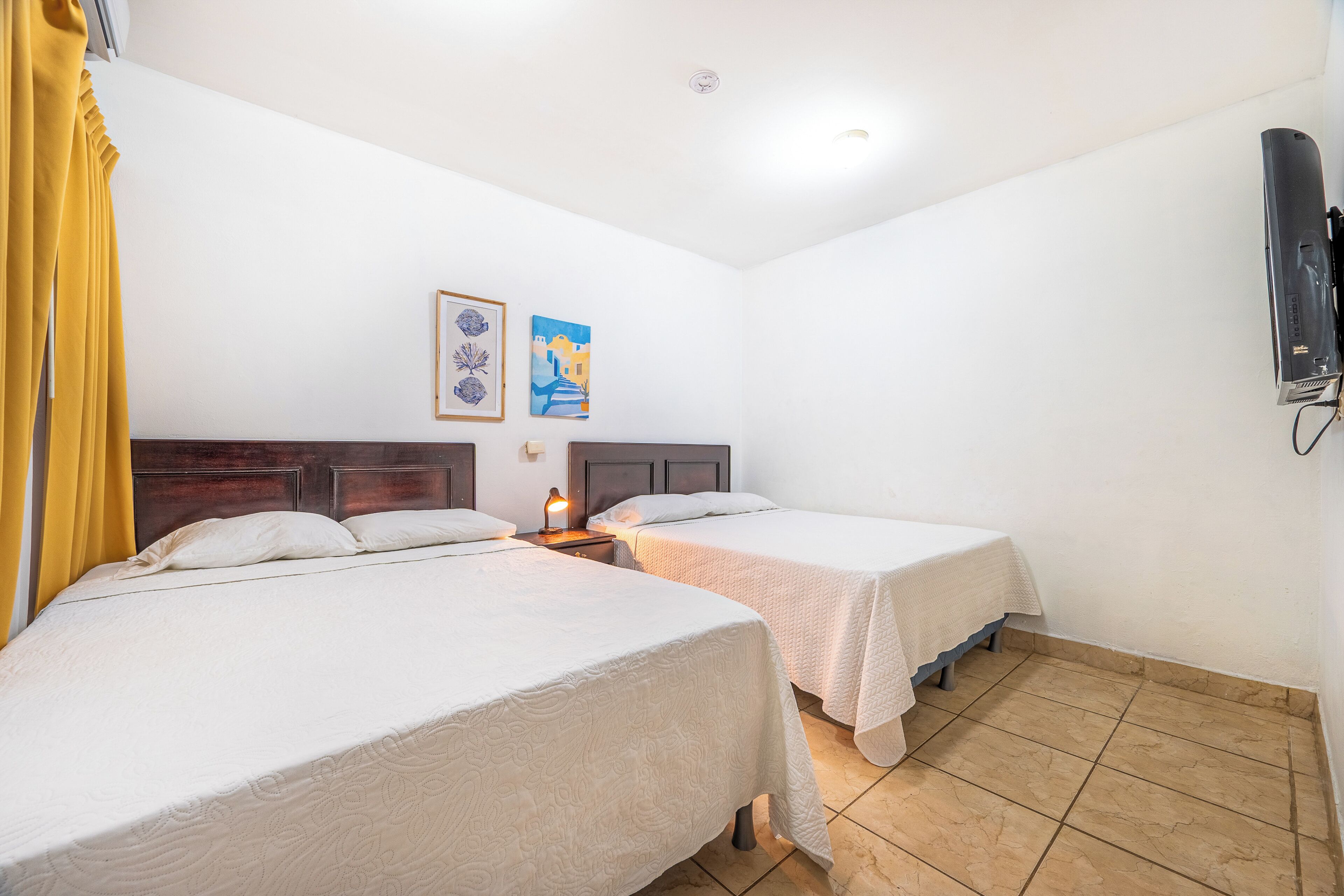 A hotel room with two beds, a wooden headboard, a flat-screen TV mounted on the wall, and a small table with a lamp.