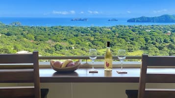 Una mesa con una botella de vino, dos copas de vino y un cuenco de fruta. La mesa está frente a un paisaje verde exuberante y el mar.