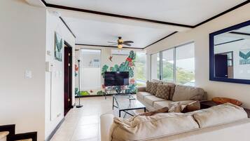 Una sala de estar moderna con un sofá, una mesa de centro y un televisor. Cuenta con un ventilador de techo y un ventanal con vistas al exterior.