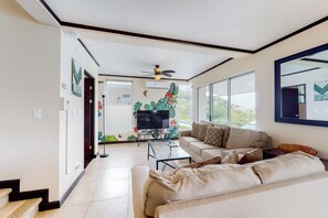 A modern living room with a sofa, a coffee table, and a TV. There is a ceiling fan and a large window with a view of the outdoors.
