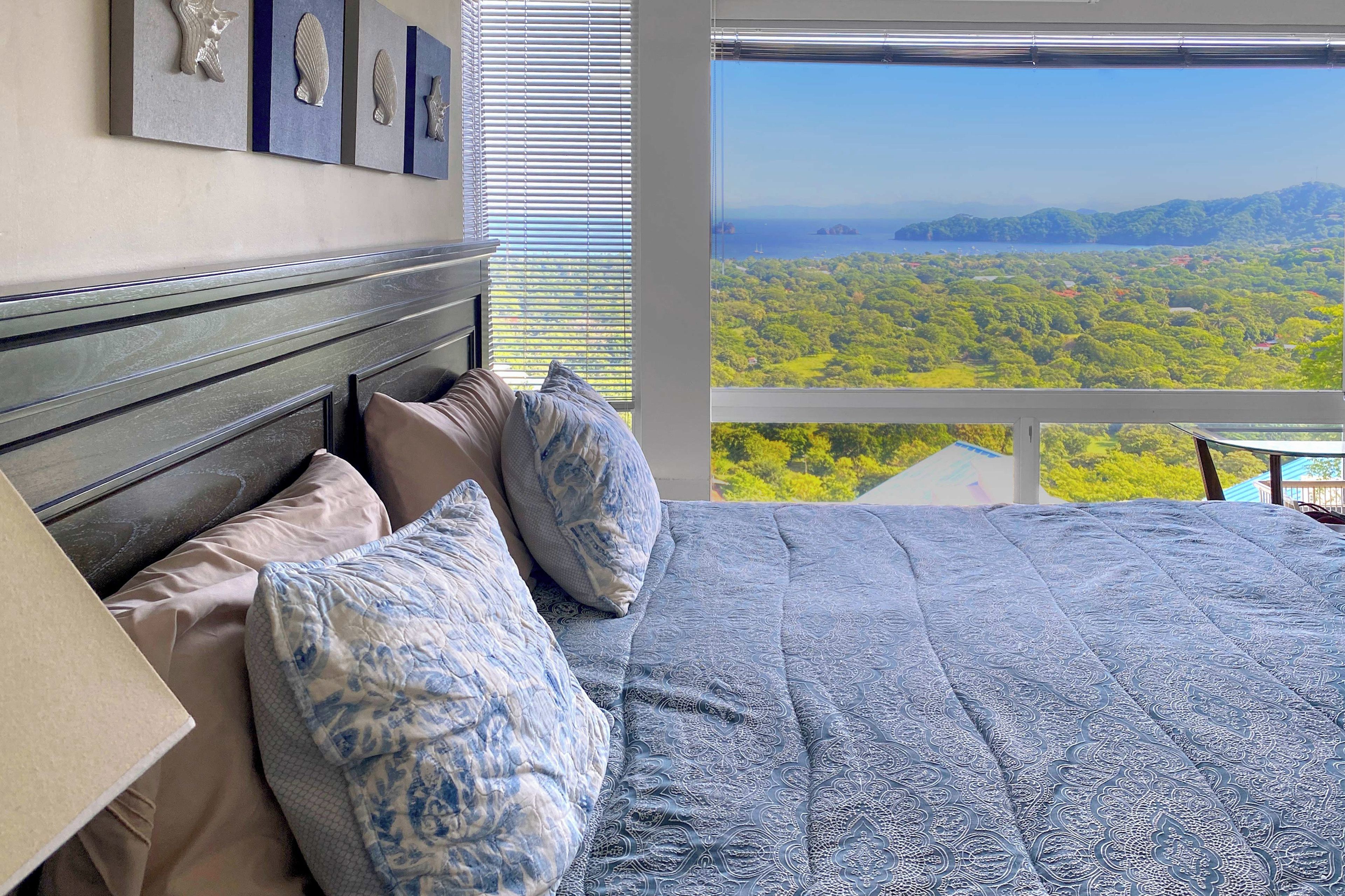 A bedroom with a large bed, multiple pillows, a headboard, and a view of a forest through a window.