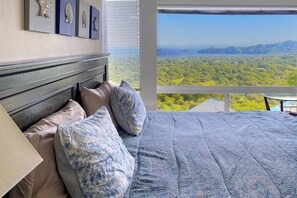 A bedroom with a large bed, multiple pillows, a headboard, and a view of a forest through a window. - Playas del Coco Home w/ Ocean-views, Deck, Pool (Sardinal)