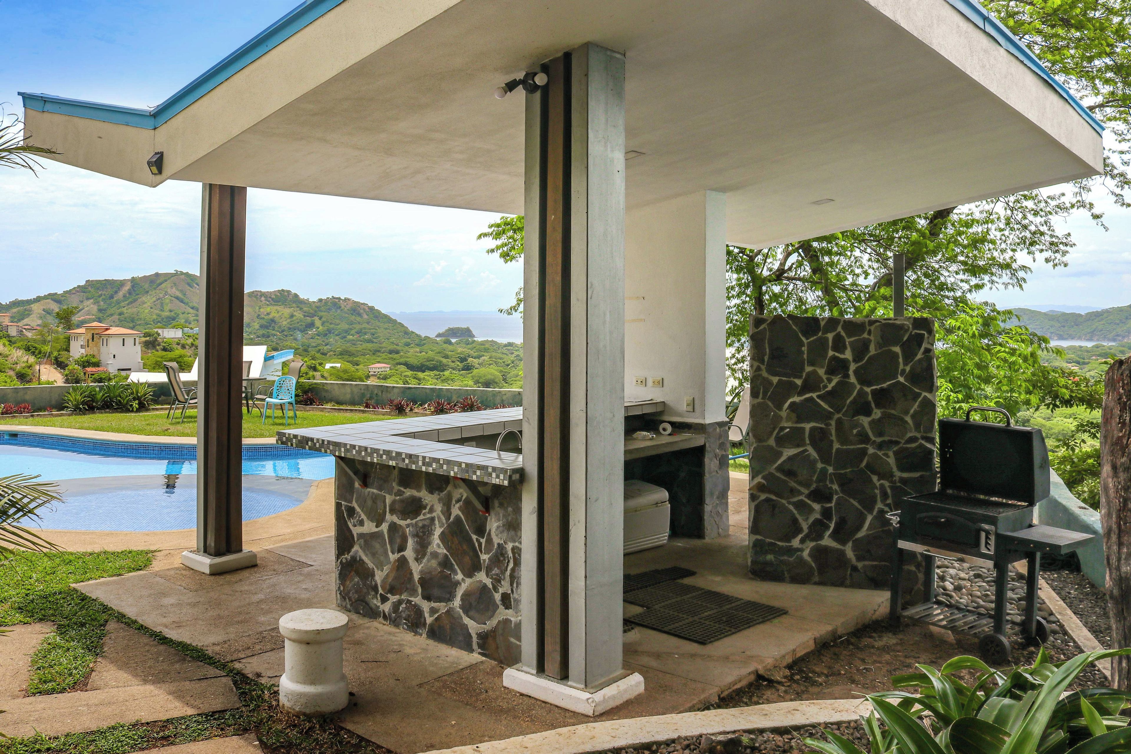 A covered outdoor area with a pool, stone barbecue, and a view of hills.
