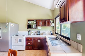 A compact kitchen with a refrigerator, stove, and sink, wooden cabinets, and a window providing a view of palm trees. - Playa Ocotal Condo w/ Pool: Walk to the Beach! (Sardinal)