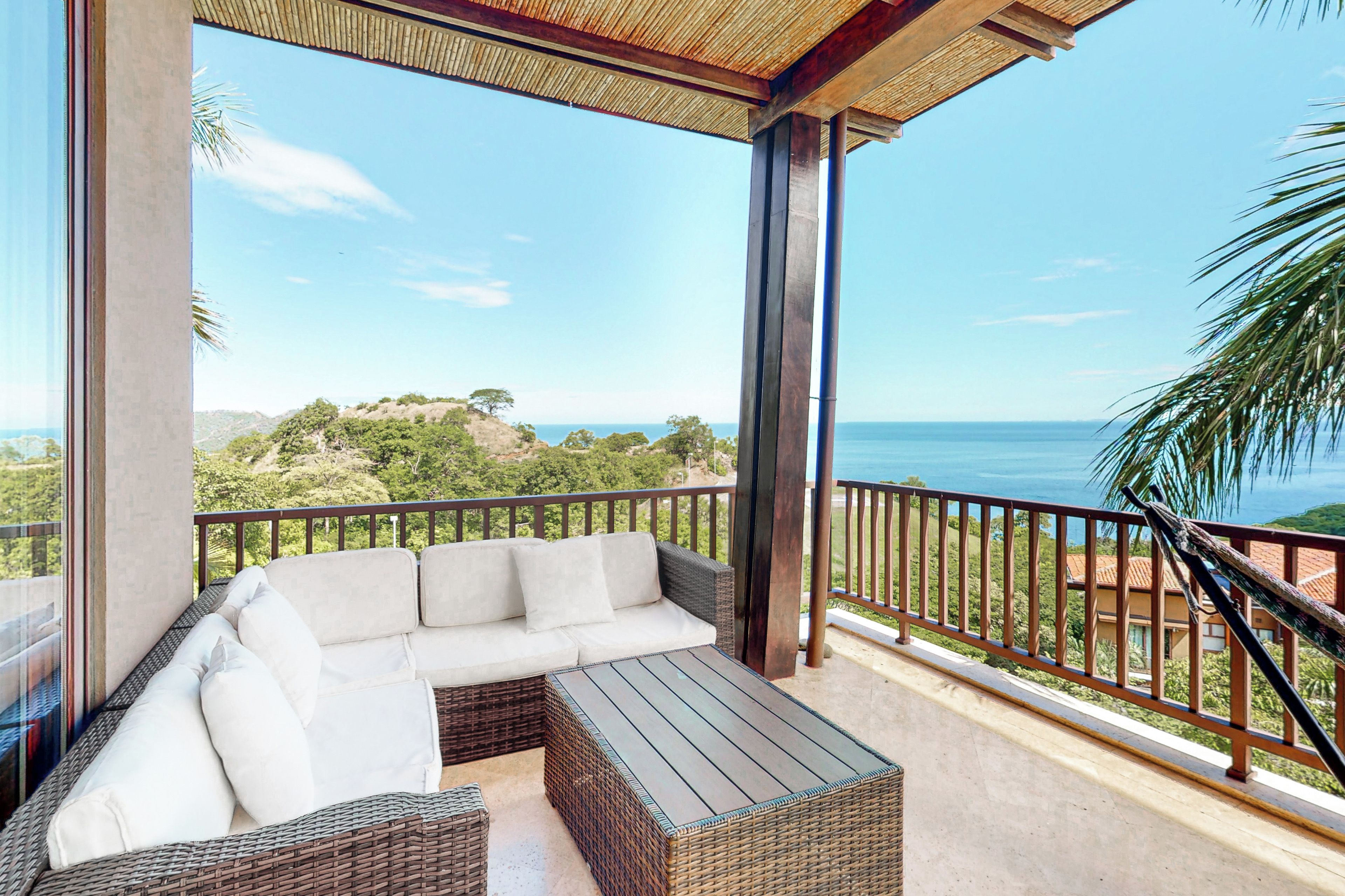 Ein Balkon mit einer Rattan-Couch und einem Tisch, mit Blick auf eine Küstenlandschaft mit Bäumen und Meer.