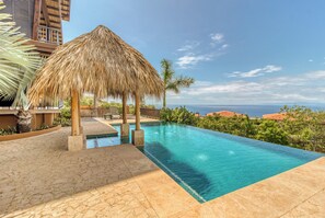 Ein Swimmingpool mit einer strohgedeckten Struktur, umgeben von Palmen und Blick auf den Ozean.