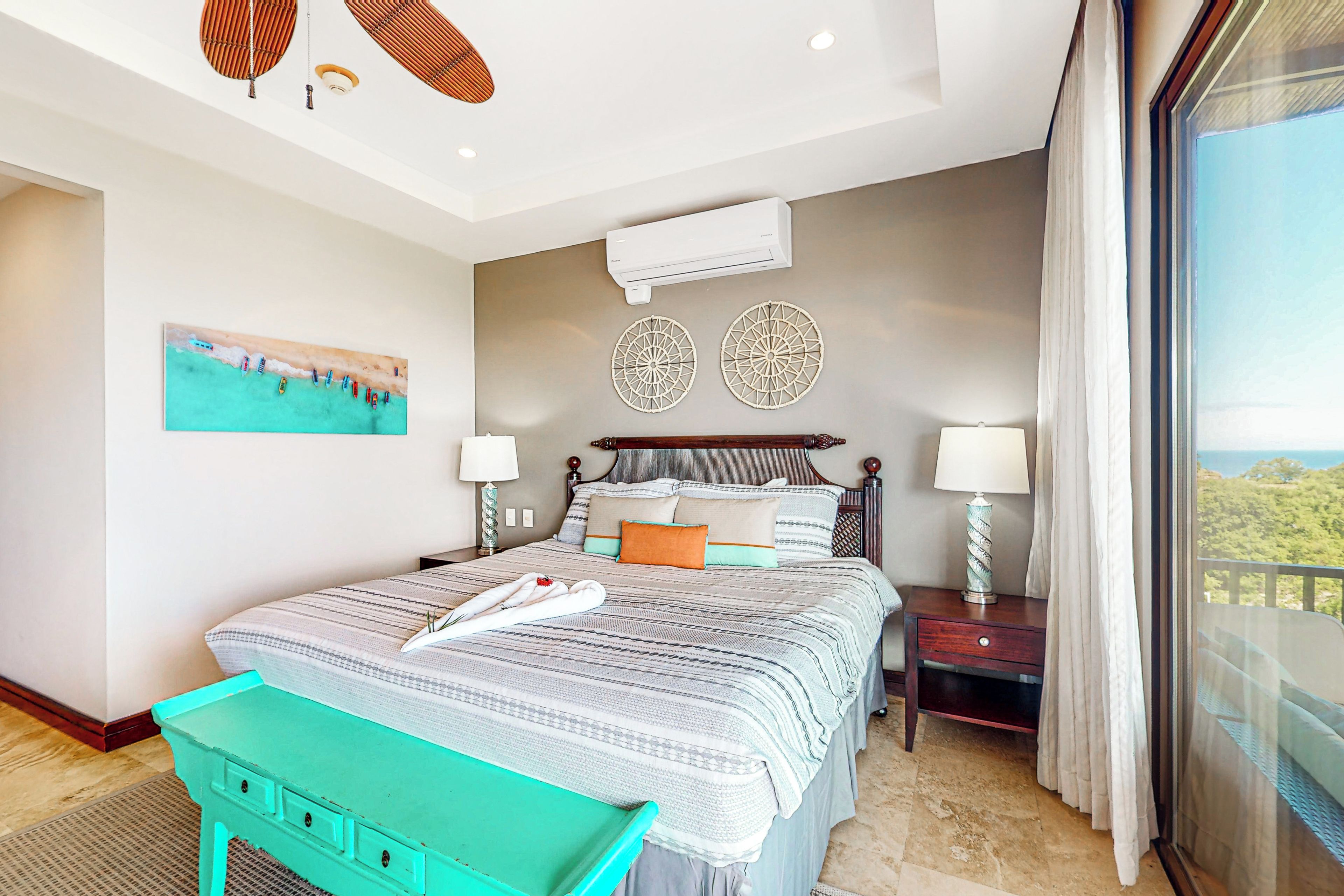 A hotel room with a bed, two wall-mounted air conditioners, a painting, and a view through a window.