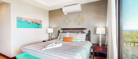 A hotel room with a bed, two wall-mounted air conditioners, a painting, and a view through a window.