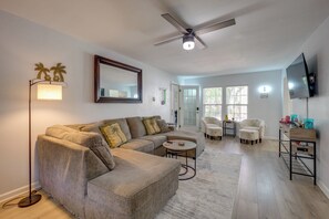 Uma sala de estar com sofá seccional, ventilador de teto, TV de tela plana e um espaço de jantar com cadeiras e mesa.