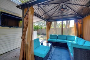 A covered outdoor seating area with a sofa and cushions, a ceiling fan, and a window overlooking a backyard. - 1 Mi to Beach! Family-friendly Waveland Home (Waveland)