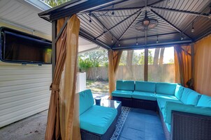 A covered outdoor seating area with a sofa and cushions, a ceiling fan, and a window overlooking a backyard.