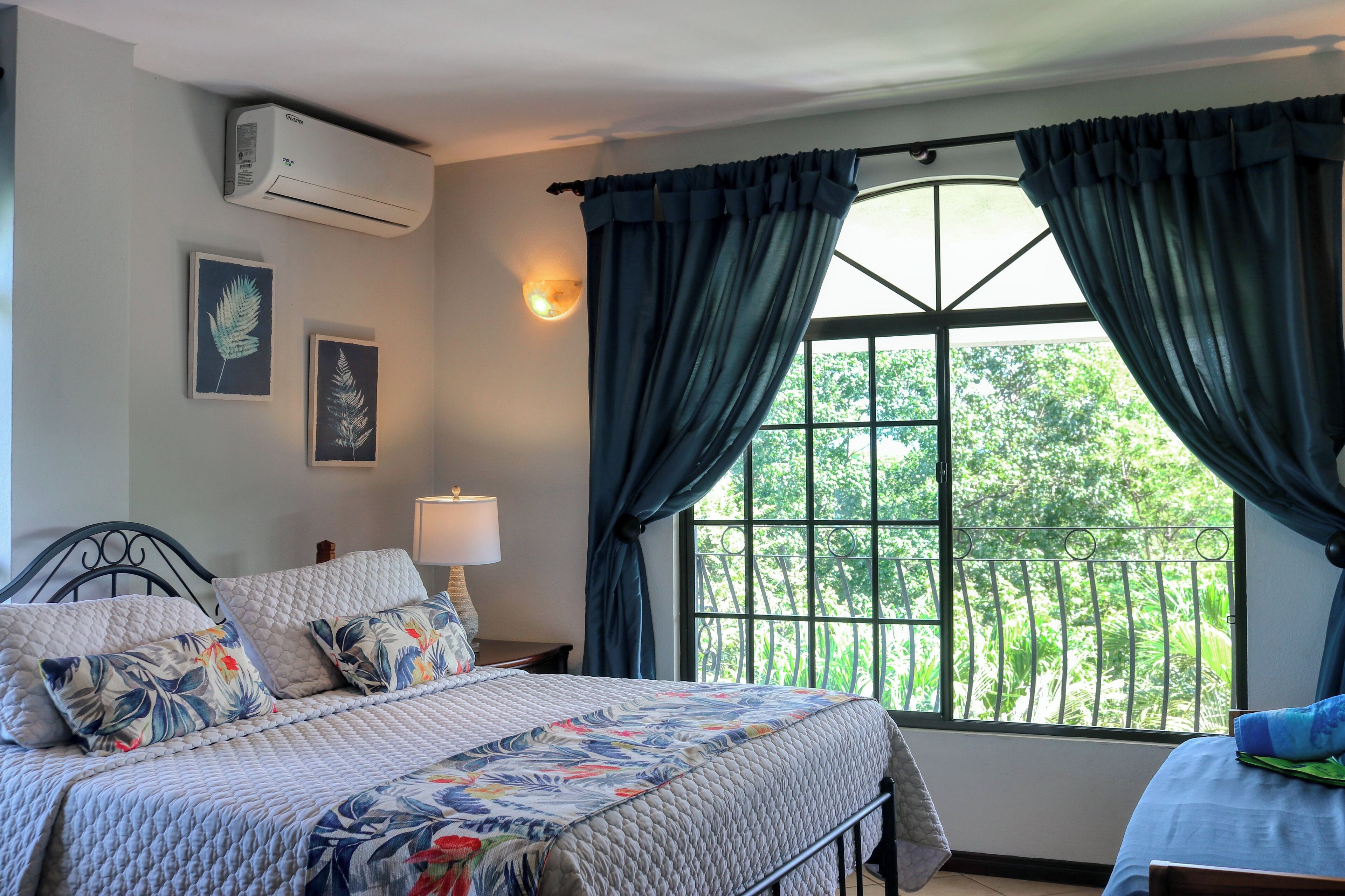 A bedroom with a bed, a nightstand, a lamp, a window with curtains, and wall-mounted air conditioning.