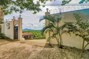 A gated entrance with a mountain backdrop, a building with a red-tiled roof, and a palm tree in the foreground. - Shared Pool & Outdoor Kitchen: Flamingo Beach Gem (Cabo Velas)