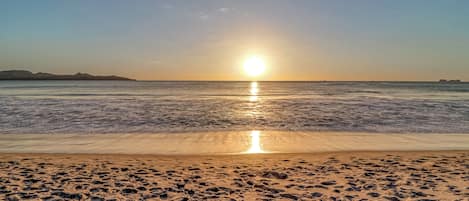 A beach with footprints leading to the water, a clear sky, and the sun reflecting on the water.