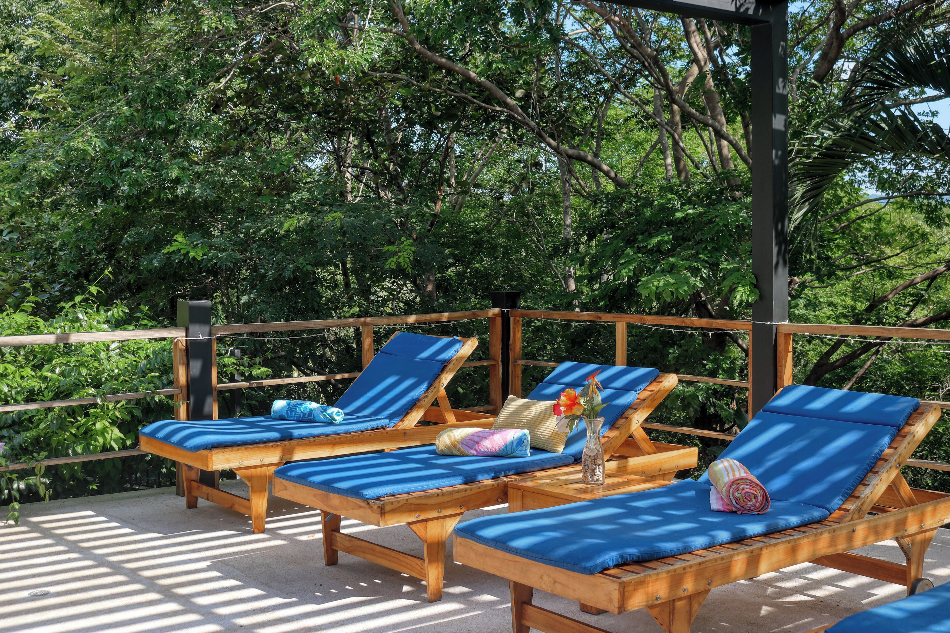 A wooden deck with three lounge chairs, each with a blue cushion, and a small table with a vase of flowers.