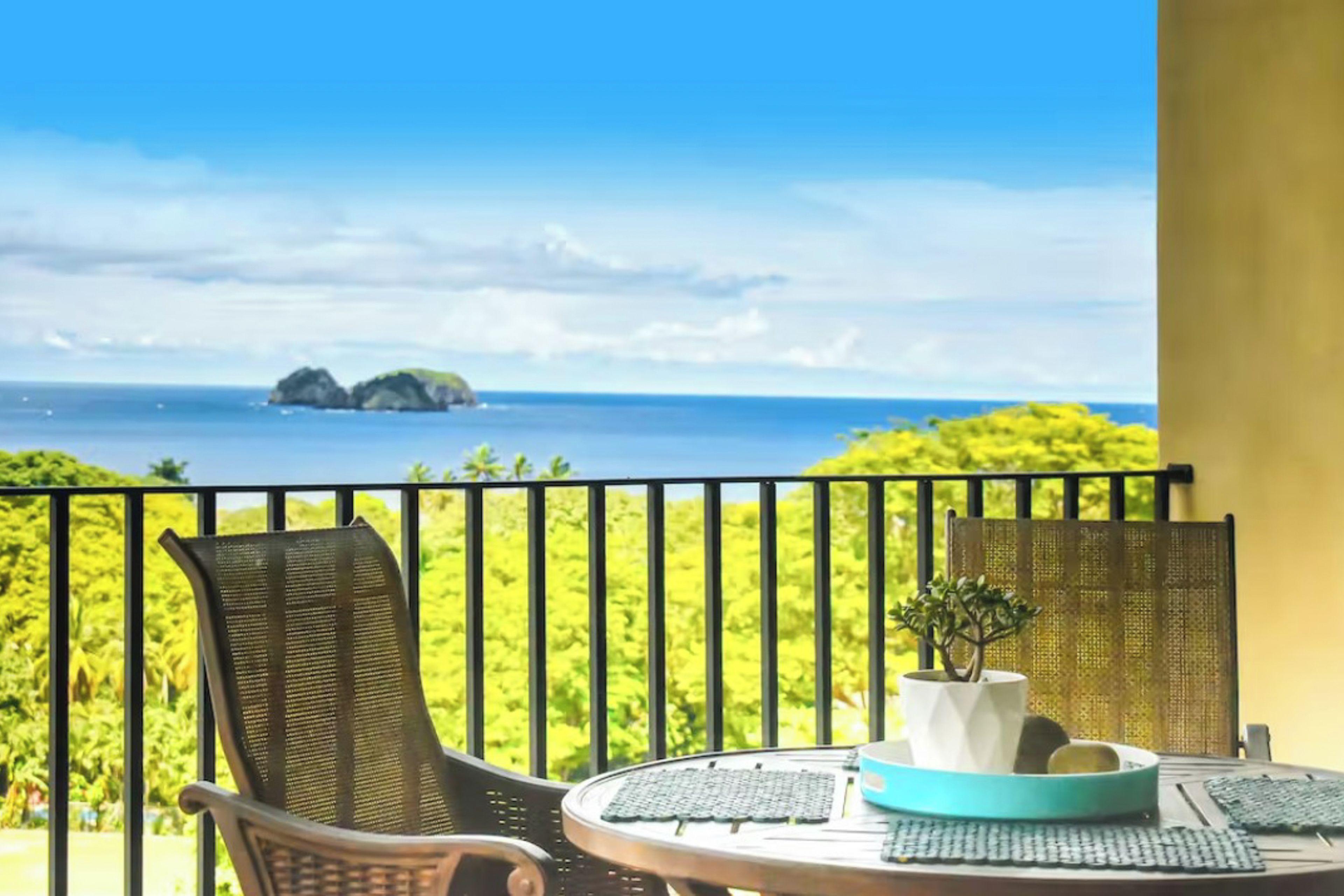 A balcony with a view of the sea and islands, a table with a plant, and a chair.