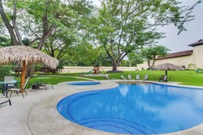 A swimming pool with a curved edge, surrounded by a grassy area with trees and a thatched umbrella. - Playa Hermosa Penthouse: Ocean Views, 2 Pools (Sardinal)