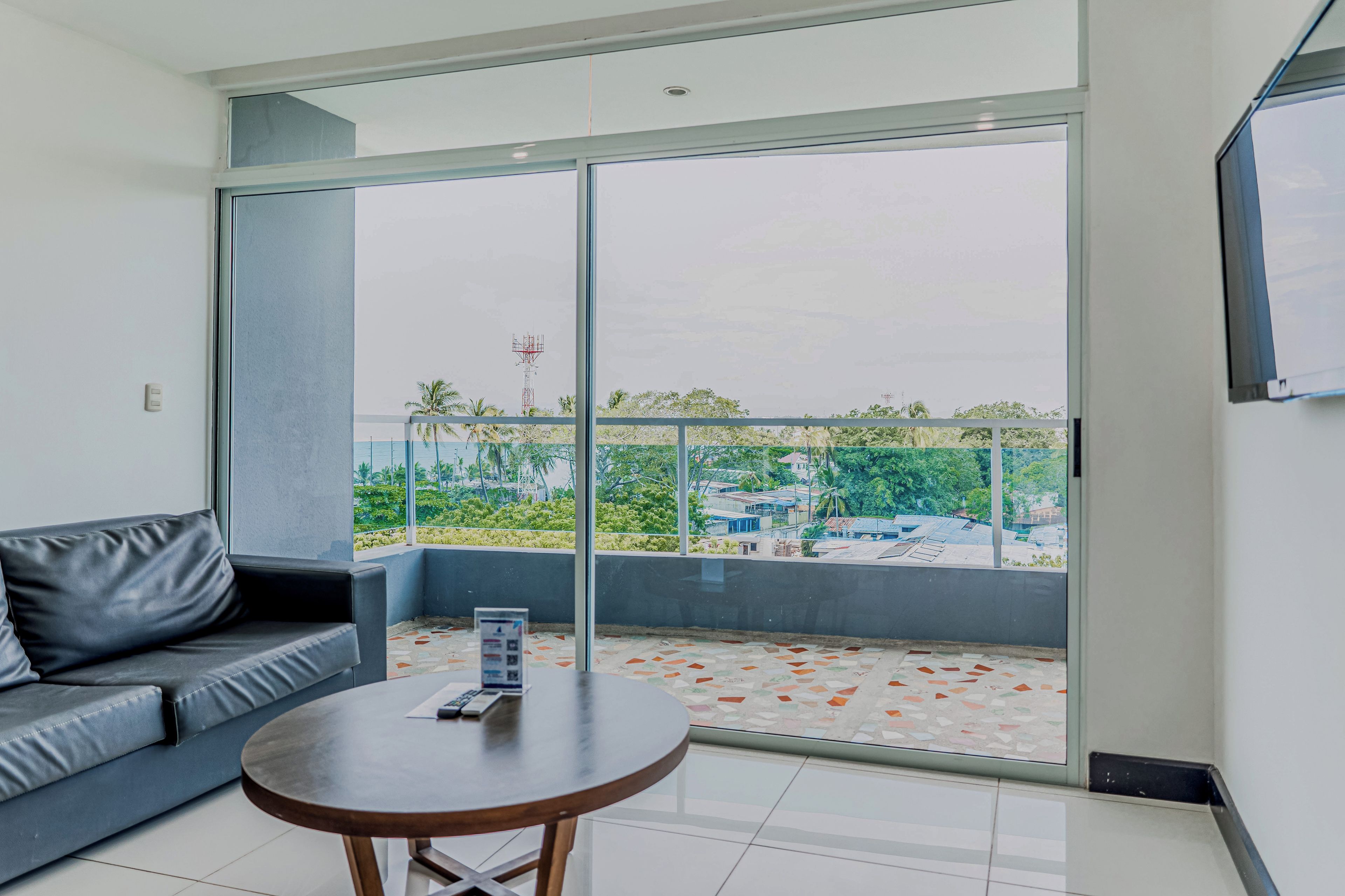 A modern living room with a grey sofa, a round wooden table, and a large sliding glass door leading to a balcony with a view of a residential area.