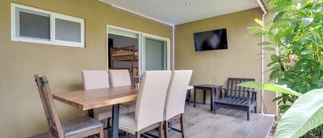 A dining area with a wooden table and chairs, a television mounted on the wall, and a window with a view of the outdoors.