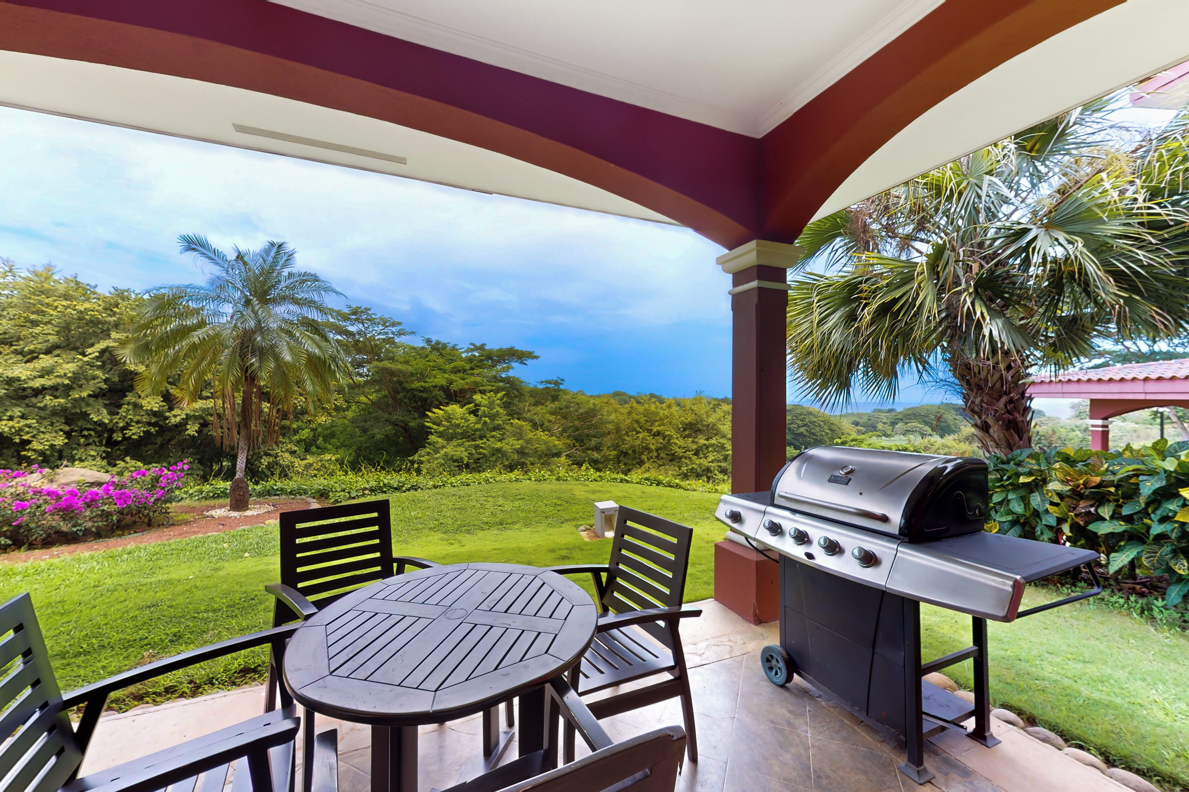An outdoor patio with a grill, seating, and a view of a lush garden.
