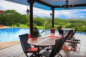 Un espacio exterior techado con piscina, mobiliario y vistas a un paisaje verde.