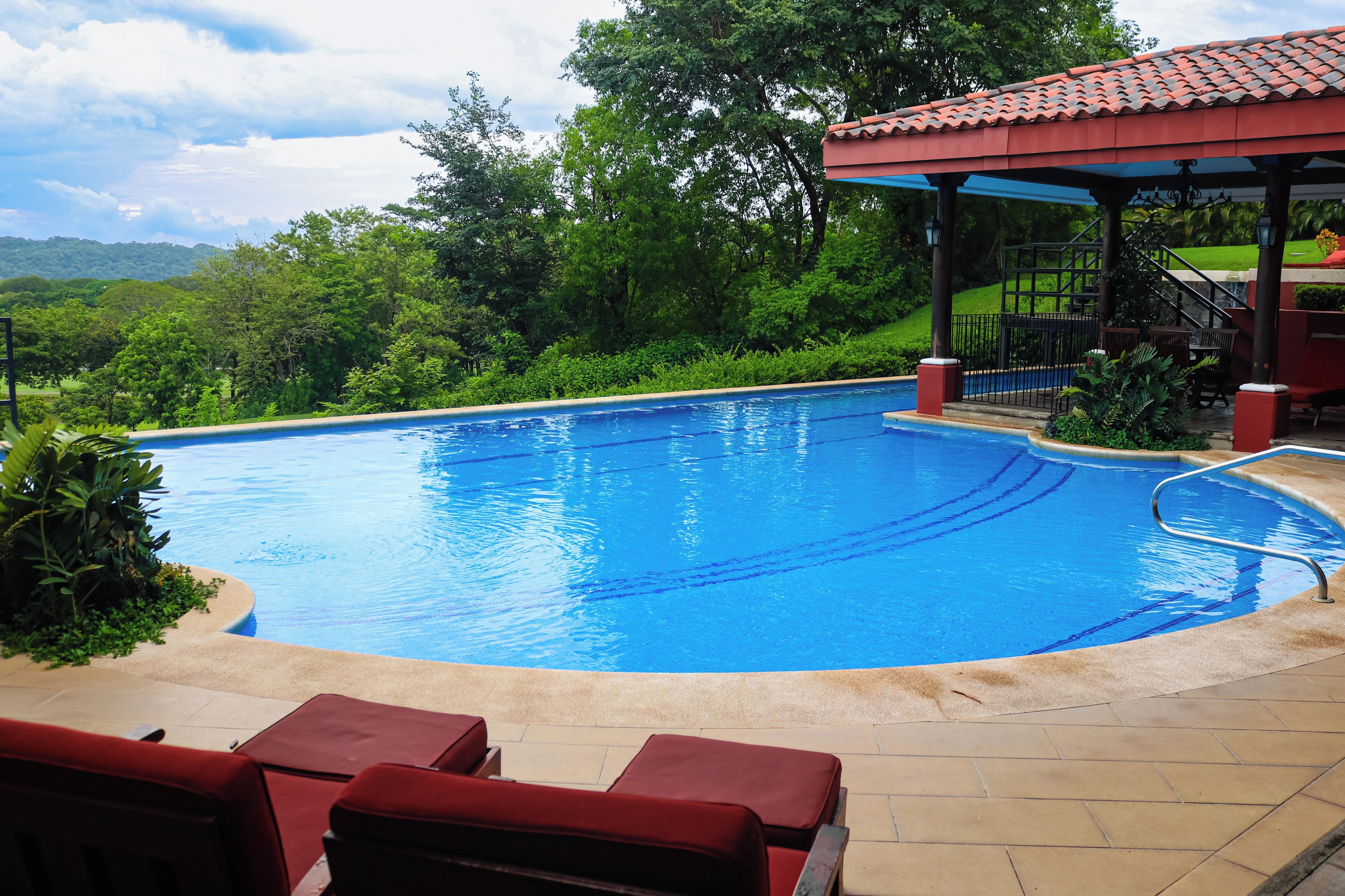 A swimming pool with a covered area, surrounded by greenery and a view of hills.