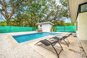 A swimming pool with two lounge chairs, surrounded by a pebble-covered deck and a green fence.