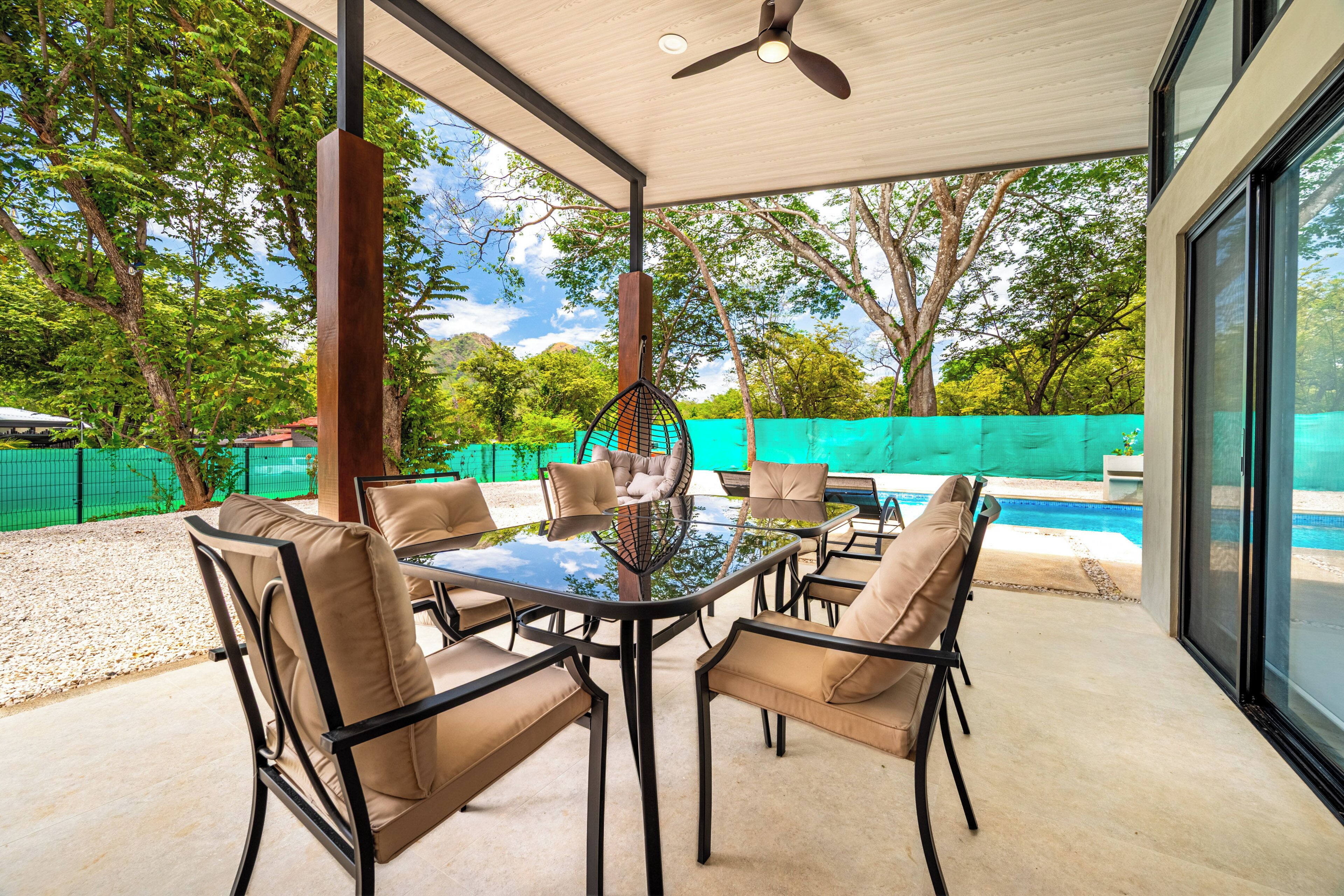 An outdoor patio with a glass-top table surrounded by chairs, a ceiling fan, and a view of a swimming pool.