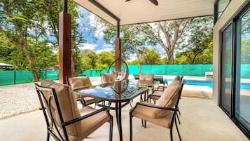 An outdoor patio with a glass-top table surrounded by chairs, a ceiling fan, and a view of a swimming pool.