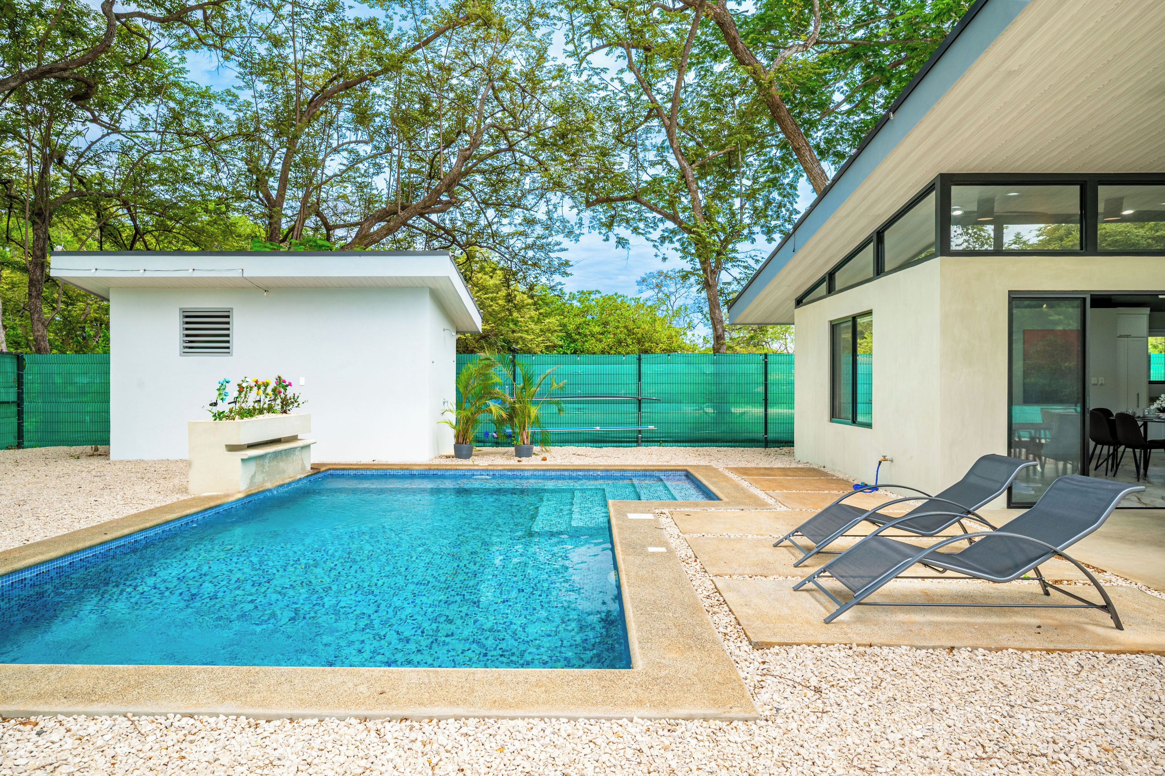 A swimming pool with lounge chairs and a small shed, surrounded by a green fence and trees.