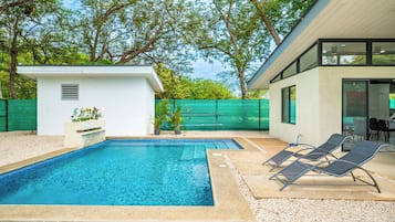 A swimming pool with lounge chairs and a small shed, surrounded by a green fence and trees.