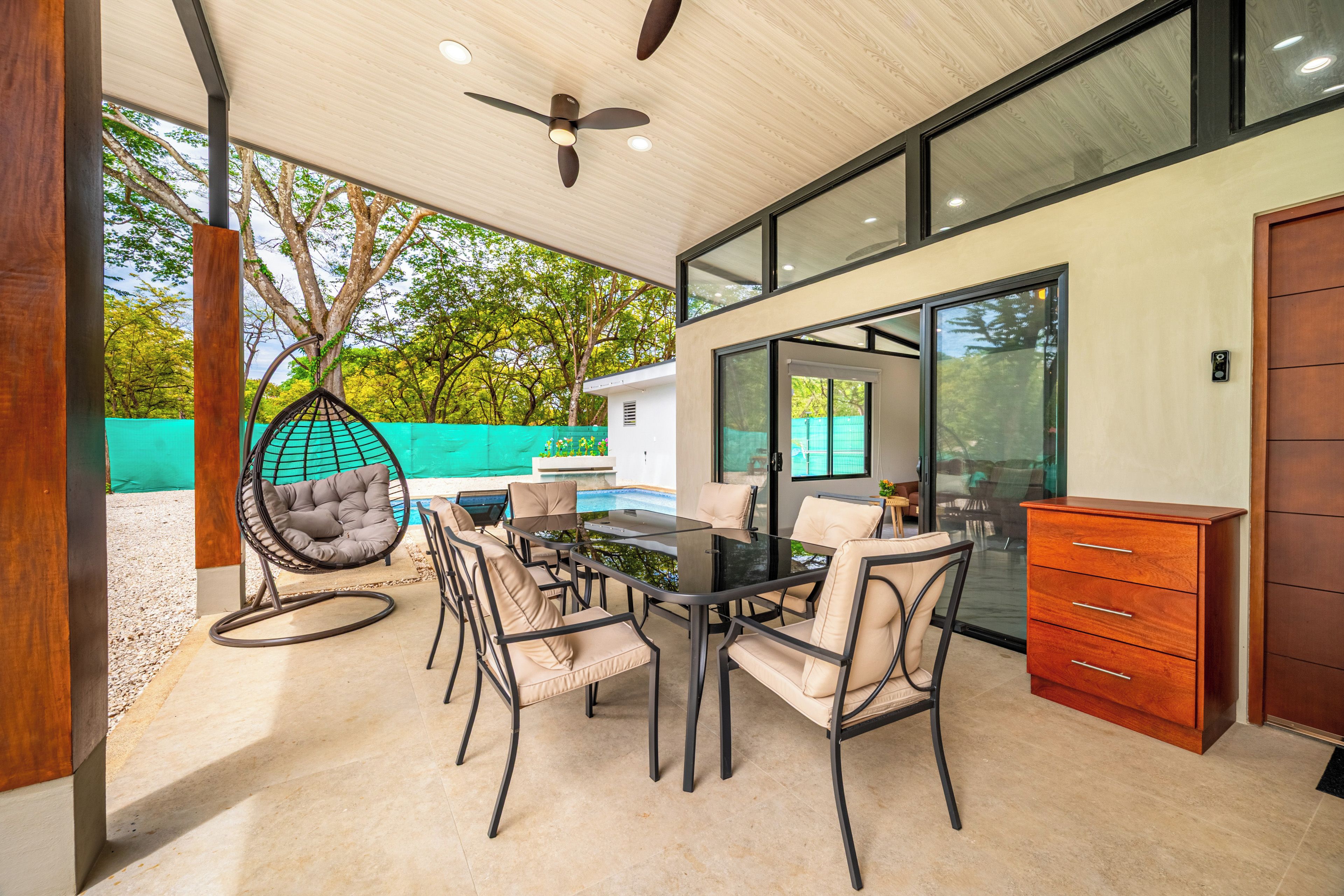 A modern outdoor dining area with a glass table and chairs, a hanging chair, and a wooden cabinet.