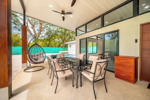 Un espacio moderno para comer al aire libre con mesa y sillas de vidrio, una silla colgante y un mueble de madera.