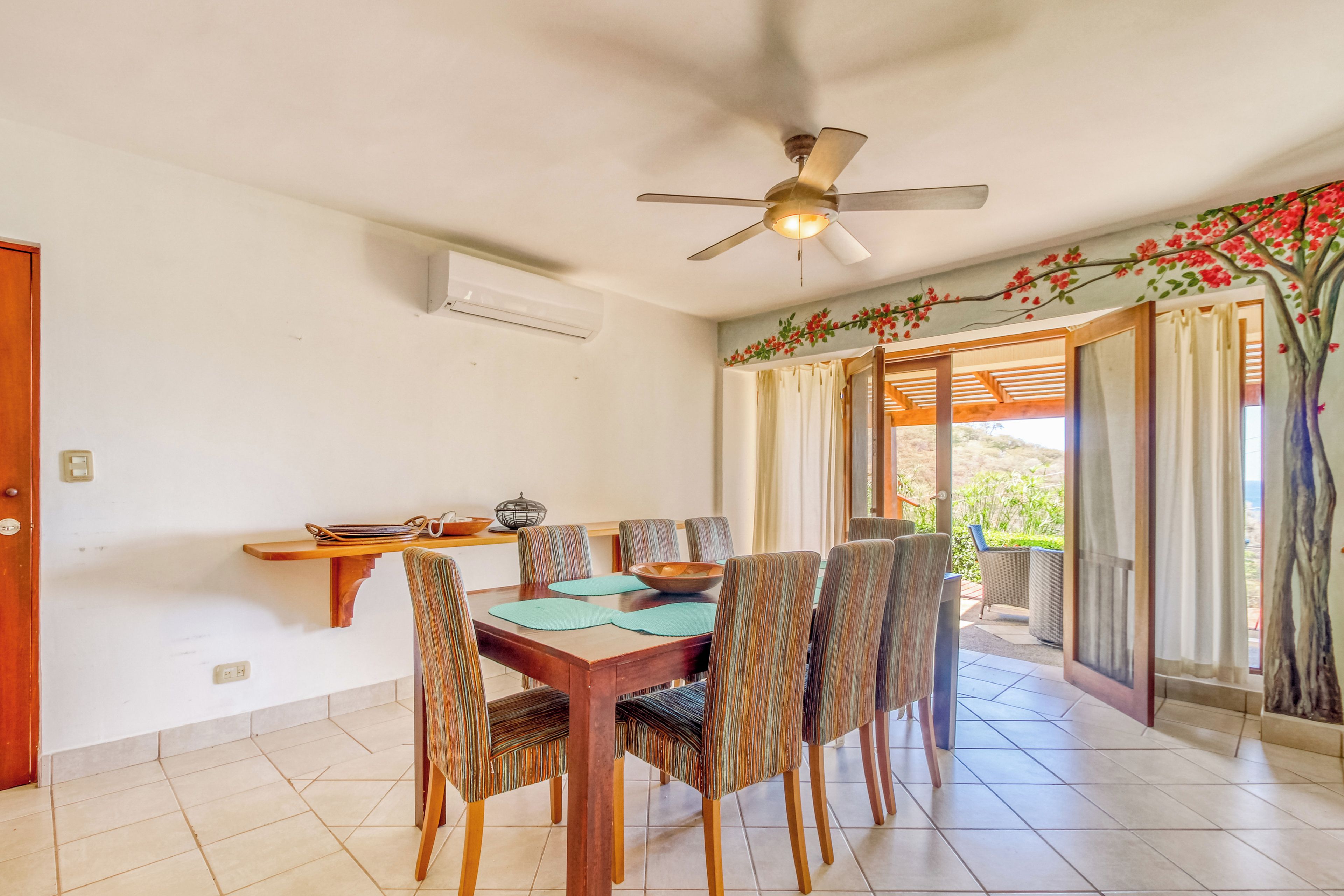 Uma sala de jantar com mesa e cadeiras de madeira, ventoinha no teto e vista através de uma porta de vidro.