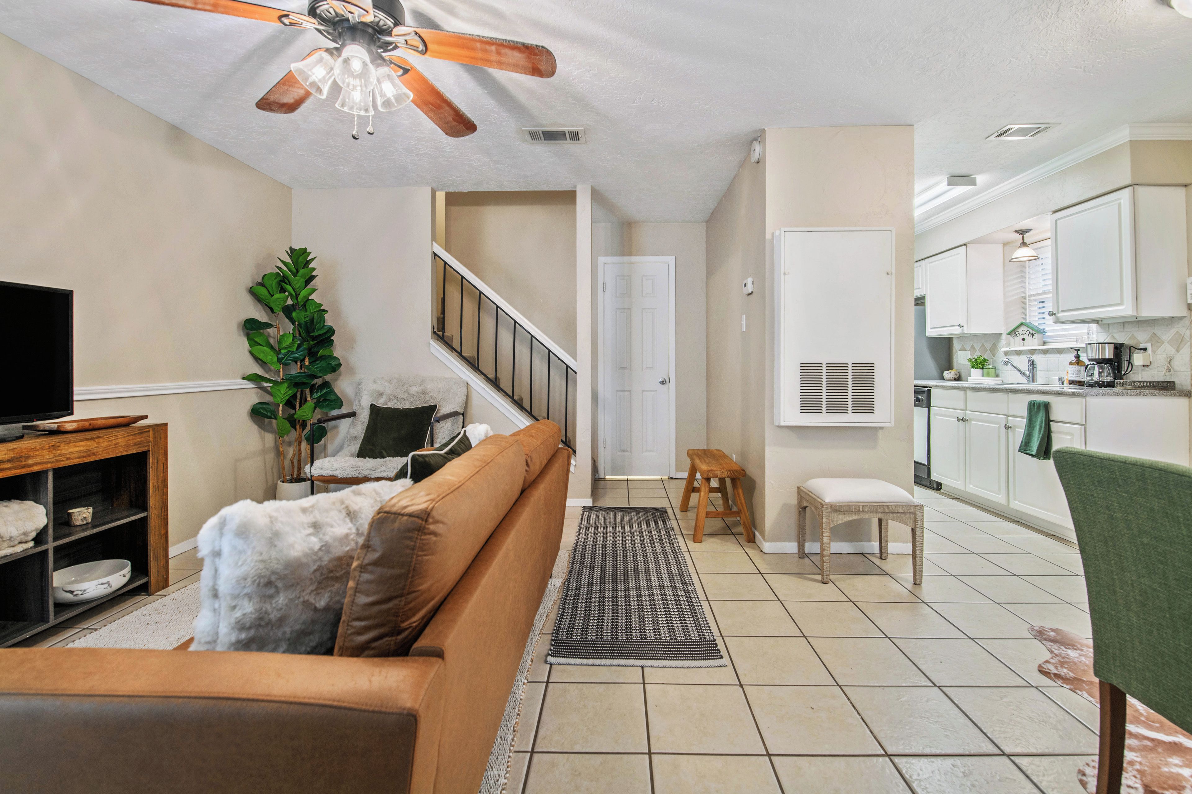 Un salon comprenant un canapé, un téléviseur, un ventilateur de plafond, un escalier et un coin cuisine avec des placards et un évier.