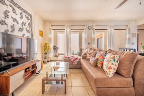 A modern living room with a flat-screen TV, a glass coffee table, a sofa with colorful pillows, and a patterned tapestry on the wall.
