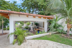 A covered outdoor area with a grill and seating, surrounded by greenery and a palm tree. - Stunning 2BR Oceanview 4th-floor Balcony (Cabo Velas)
