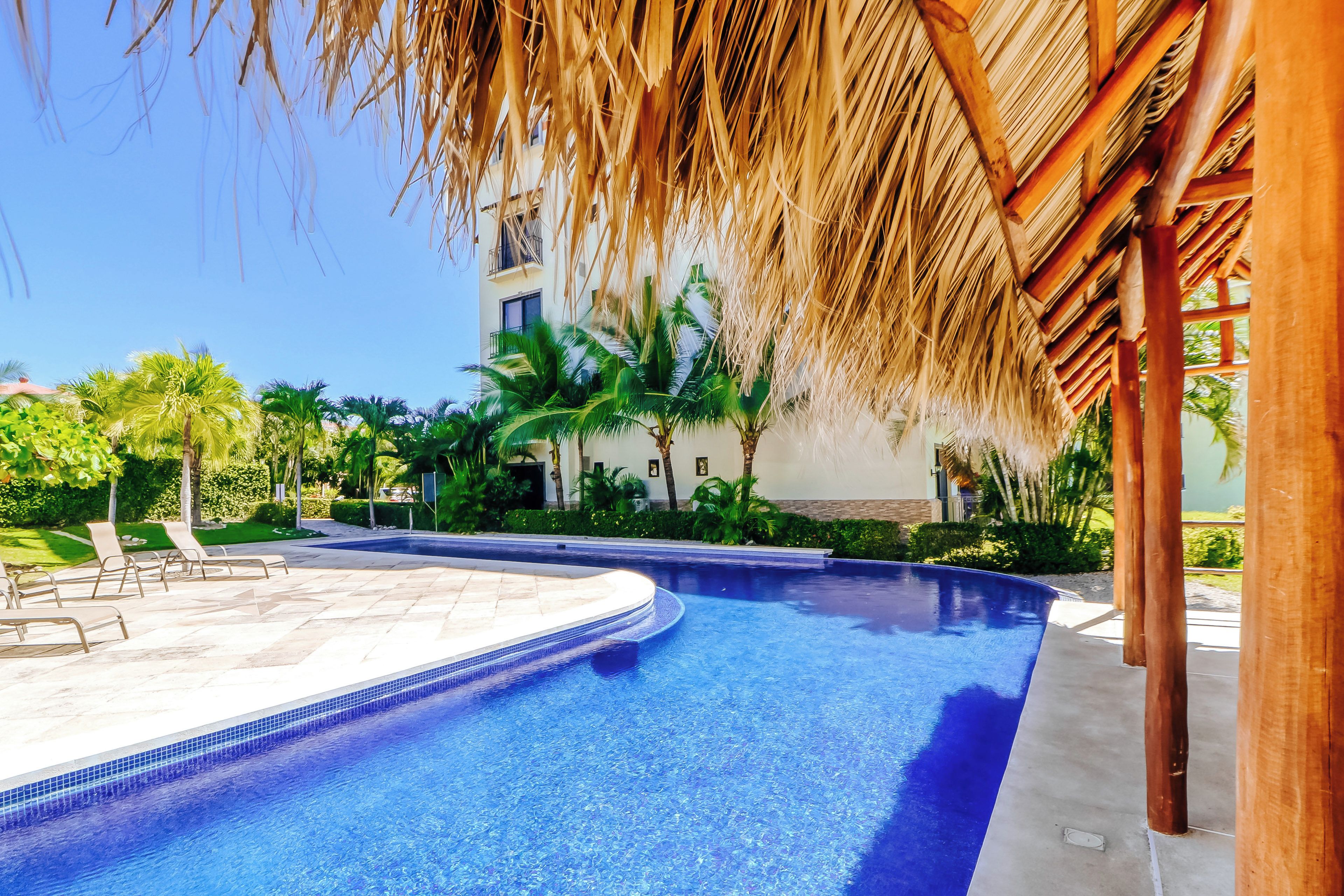 A swimming pool with a clear blue water, surrounded by palm trees and a building with a thatched roof.