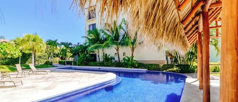 A swimming pool with a clear blue water, surrounded by palm trees and a building with a thatched roof.
