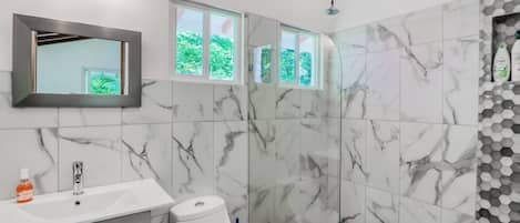 A bathroom with a white sink, a toilet, and a shower area with a glass door. There are windows above the shower, and a mirror above the sink.